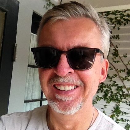 Photograph of Christopher Looby, wearing sunglasses and standing in front of a white wall with vines growing on it.