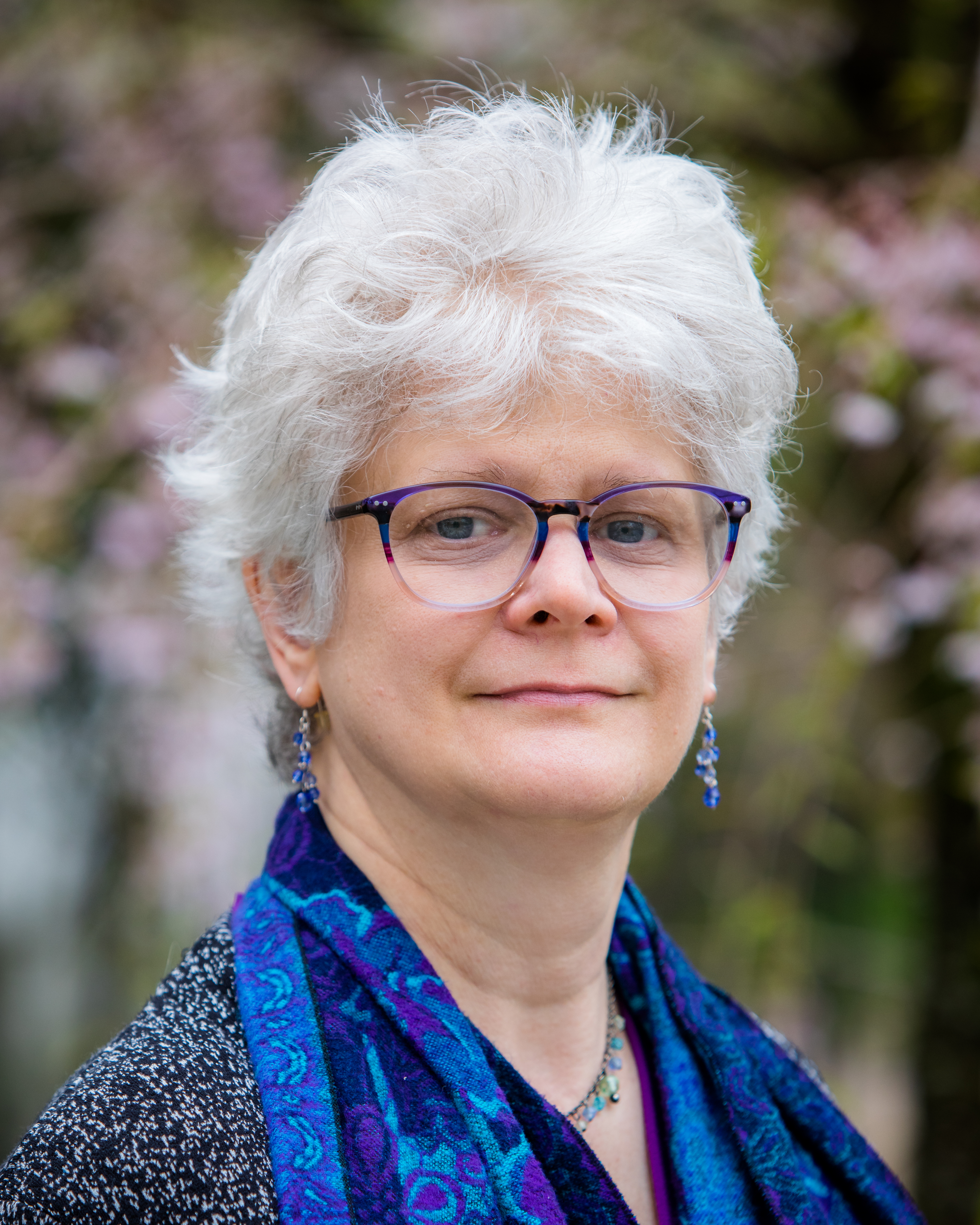Photo of Laura Wasowicz with short white hair wearing glasses and a blue-purple scarf, standing in front of a blurred floral background.