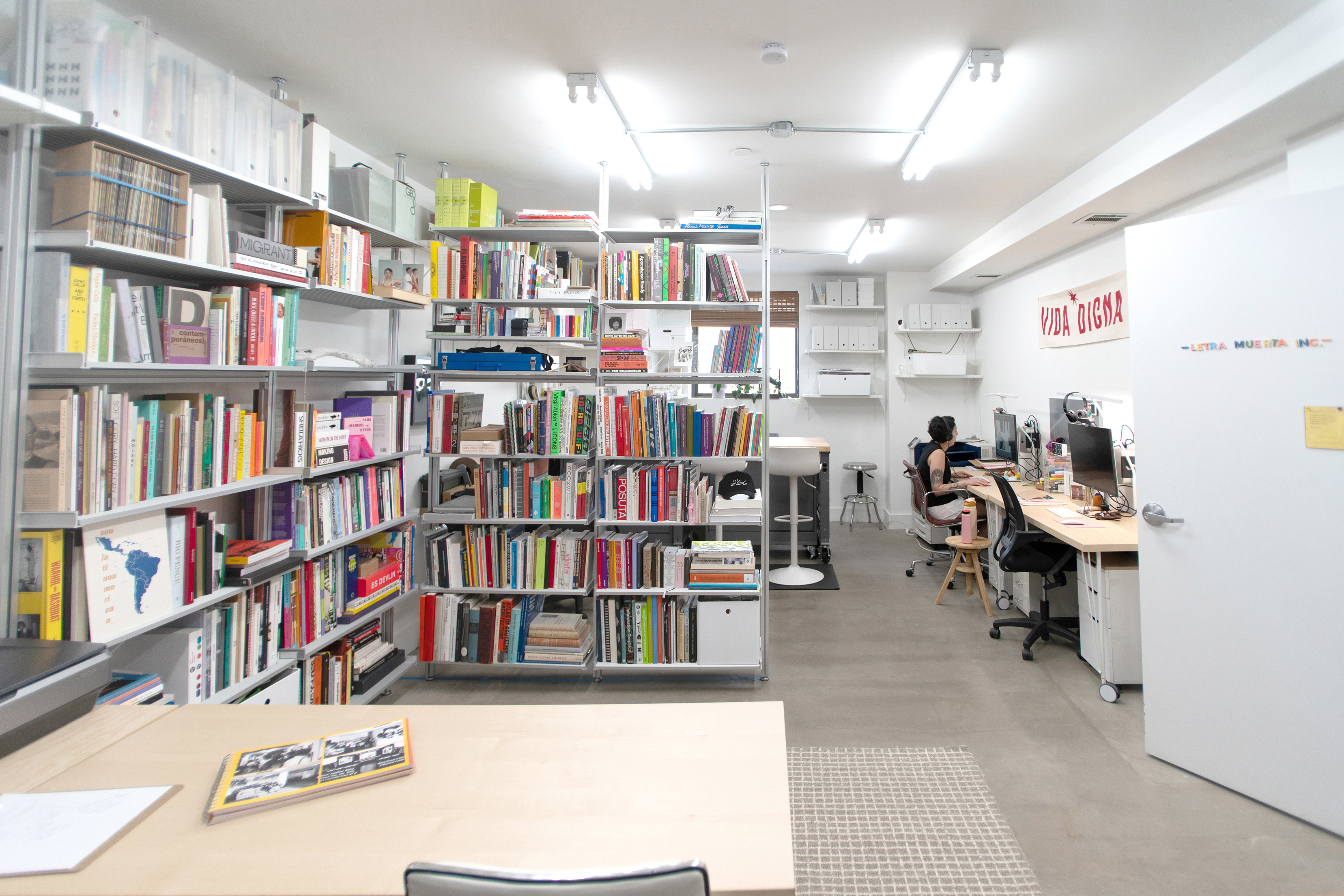 Bright library office with shelves of colorful books and a person working at a desk with dual monitors. Room has a modern, organized feel.