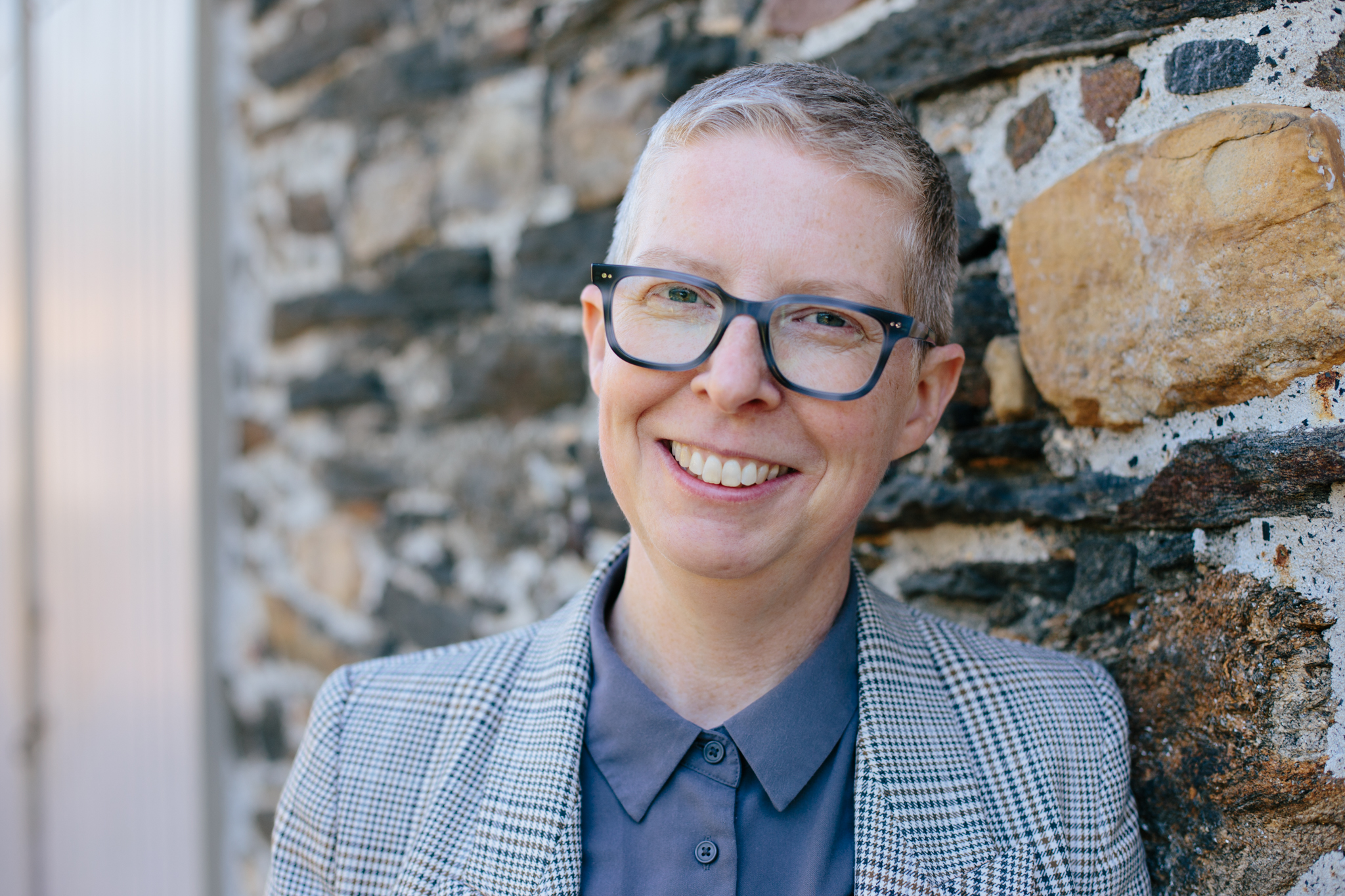 Photo of Laura Helton, with short hair, glasses, and wearing a collared shirt and blazer, smiling in front of a stone wall.