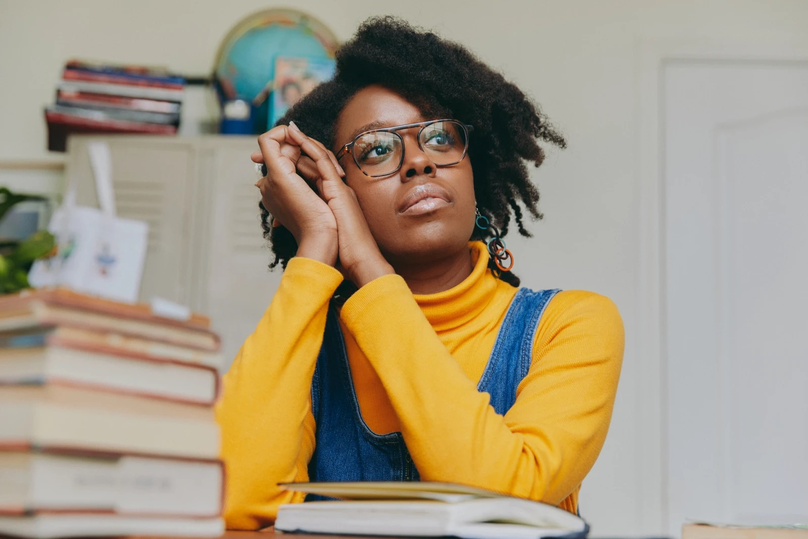 Photo of Katie Mitchell, wearing glasses and a yellow long-sleeved shirt, sitting in an office. A stack of books is on the table surface in front of her. A set of lockers with more books and a globe is behind her.